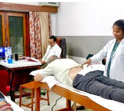 A physiotherapist in a white coat treating a patient lying on a couch while a colleague looks on from behind