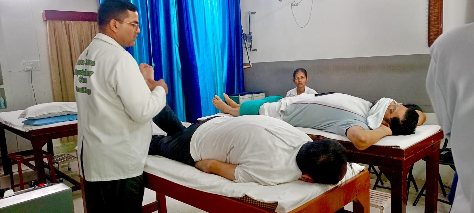 A therapist in a white coat standing beside a patient lying on a couch, with a second patient and a staff member seated behind
