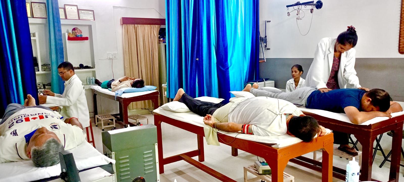 A therapist in a white coat treating a patient’s back in the treatment room, with other patients on couches beside her
